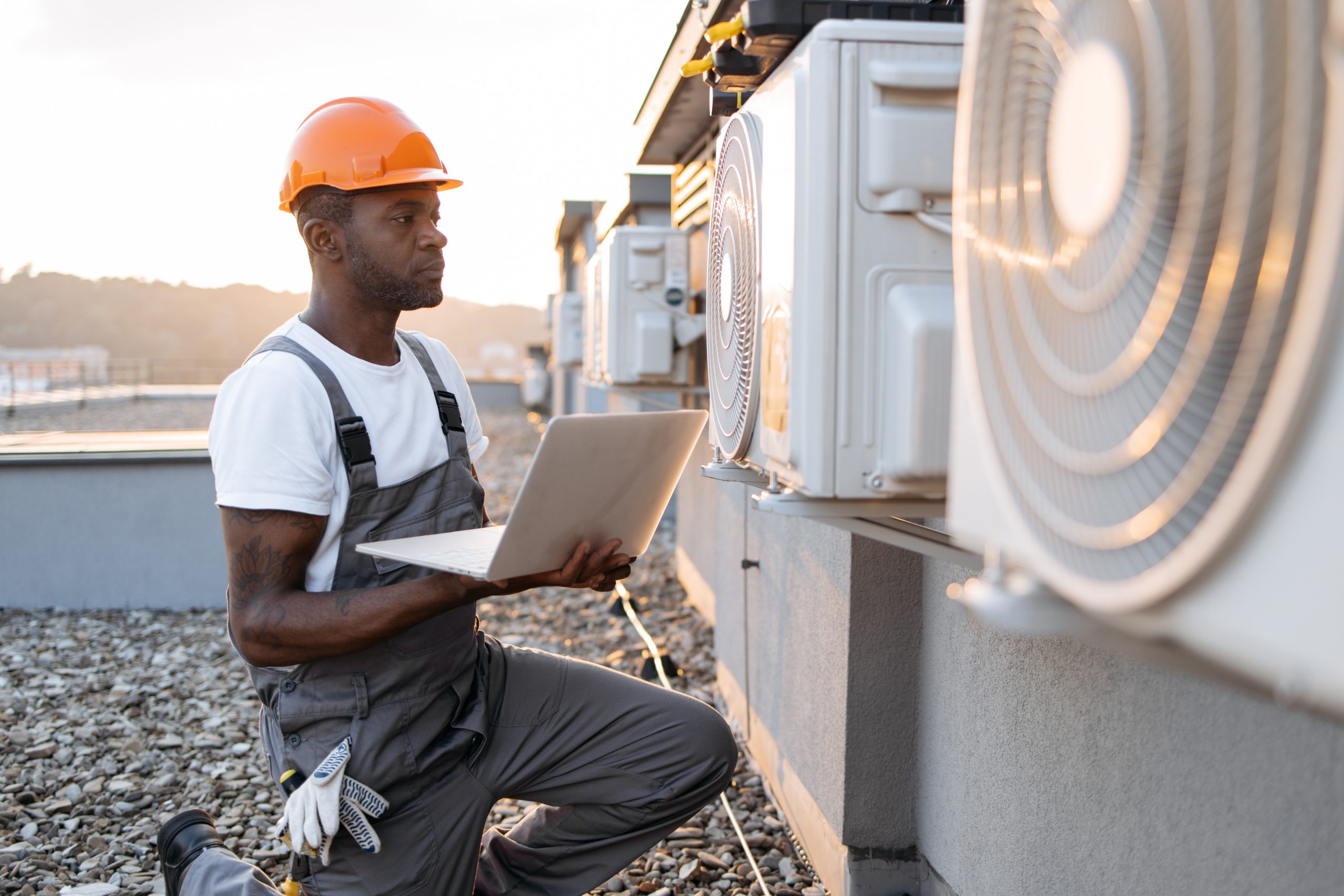 Side view of skilled man typing on wireless laptop while kneeling near hanging air conditioner outdoors. African american specialist using modern technologies for quickly repairing of cooling system.