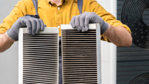 A technician wearing gray gloves holding two air conditioning filters side by side — one clean and one heavily dust-clogged — for a before-and-after comparison.