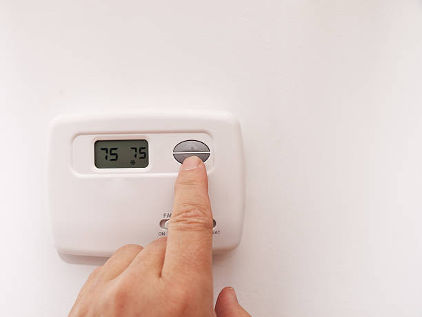 A person's hand pressing the button on a white digital wall thermostat displaying 75°F on both the current and set temperature readings.